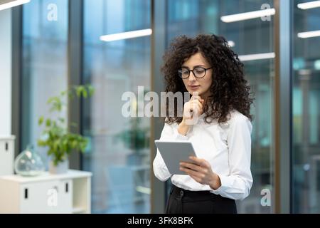 Businesswoman using a tablet for digital strategy and corporate planning in a modern office. Focused professional woman in glasses reviewing data and information for business development tasks. Stock Photo