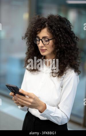 Businesswoman using smartphone for corporate communication and professional tasks. Focused woman in white shirt checking mobile phone for work updates in a modern office environment. Stock Photo