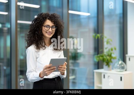 Professional businesswoman holding a tablet in a modern office, smiling female employee using digital technology for corporate communication and business tasks. Stock Photo