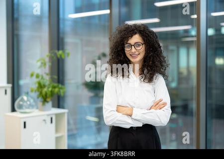 Professional businesswoman with curly hair and glasses standing in a modern office. This confident female employee is smiling with arms crossed for a corporate portrait in the workplace. Stock Photo