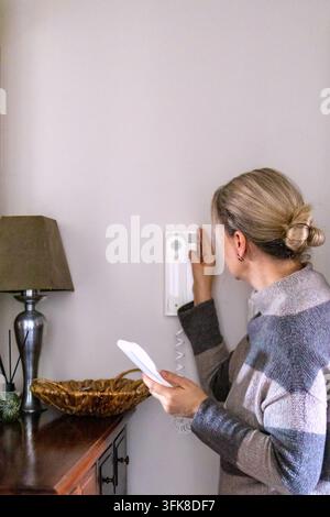 Mid adult woman holding a telephone handset while pressing a button on a wall security intercom system. Concept of apartment smart automation, residen Stock Photo