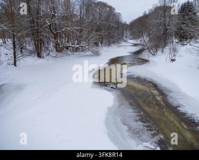 View of the partially frozen Vilnia River flowing through a snow-covered forest in Vilnius, Vilnius County, Lithuania. Stock Photo