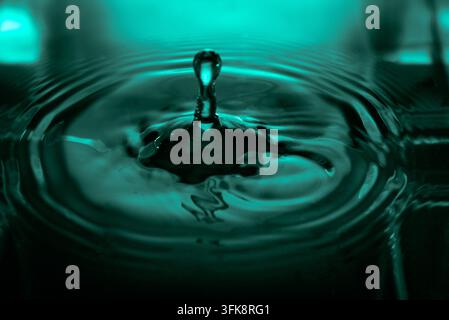 190.A vibrant macro image capturing the precise moment a water drop impacts a teal-lit surface, forming a distinct splash and concentric ripples. The Stock Photo