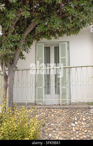A white building facade features light green shutters on a glass door, a wrought iron railing, and a large tree providing shade. A stone textured wall Stock Photo