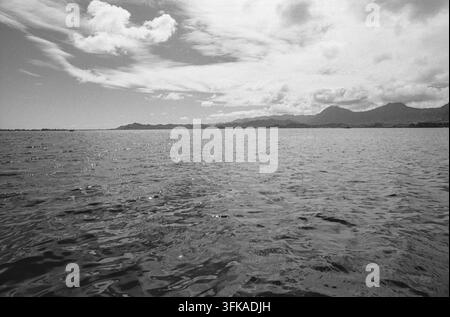 Kaneohe Bay, captured circa 1985, presents a serene coastal landscape on the island of Oahu. The expansive bay features gently rippling water extending towards the distant, verdant mountains that define the shoreline. Overcast skies create soft lighting across the scene, highlighting the natural beauty of this Hawaiian destination. Kaneohe Bay, Kaneohe, Honolulu, Oahu, Hawaii, USA Stock Photo