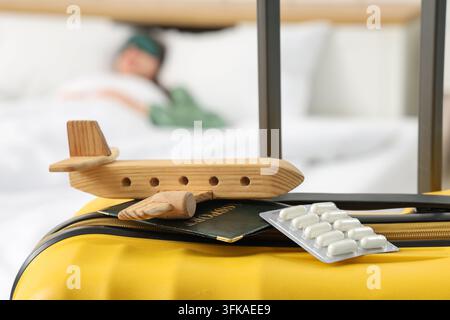 Travel sickness and health travel insurance, wooden airplane model with passport and medication pills on suitcase in hotel room with sleeping person Stock Photo