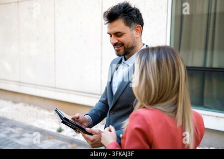 Two people are engaged in a business discussion outside a modern building. They are using a tablet. Stock Photo