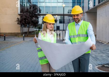 Two construction workers stand together outside a building. They look at plans and discuss their project. Stock Photo