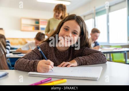 Smiling schoolgirl writing in classroom looking at camera Stock Photo
