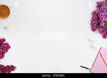 Feminine white desk with purple lilac branches, a cup of coffee, gold paper clips and a pink notebook with pen, large empty space in the center. Stock Photo