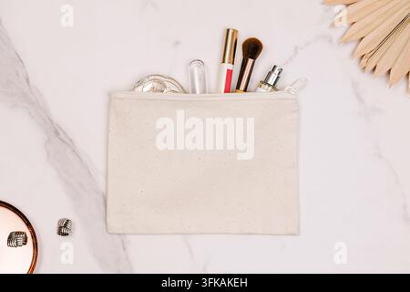 Blank natural canvas zipper pouch filled with makeup brush, lipstick, perfume and compact on white marble with dried palm leaf, top view. Stock Photo