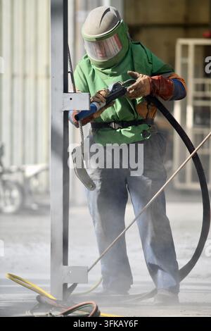 A man in protective clothing operates a sandblaster to clean steelwork on a fire-damaged building, ready for repainting. Stock Photo