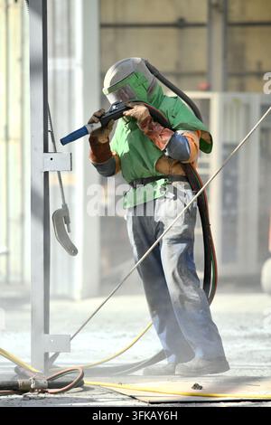 A man in protective clothing operates a sandblaster to clean steelwork on a fire-damaged building, ready for repainting. Stock Photo