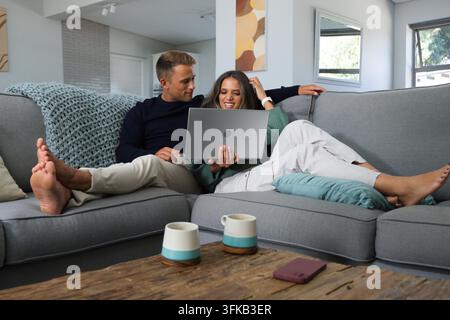 Diverse couple sitting on gray sectional sofa at home, using laptop with mugs nearby. Interior, minimalist, cozy, modern, lounge, remote, focused Stock Photo