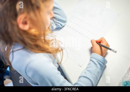 First grade girl practices handwriting with pencil in school workbook. Child learns to write letters during early education with selective focus and Stock Photo