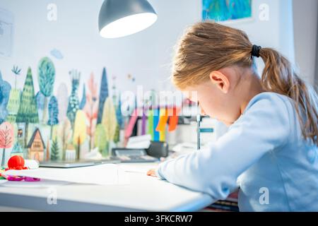 First grade girl practices handwriting at desk in her room. Child studies at home at beginning of school year with selective focus and soft background Stock Photo