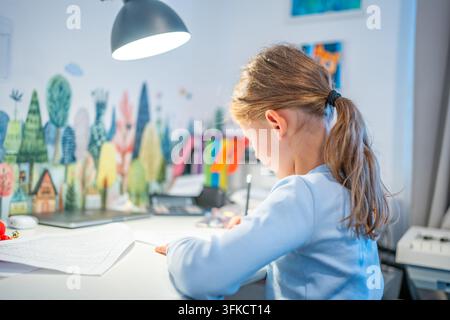 First grade girl practices handwriting at desk in her room. Child studies at home at beginning of school year with selective focus and soft background Stock Photo