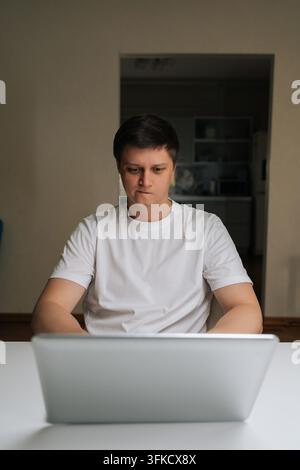Vertical portrait of young man wearing white t-shirt sitting at table, deeply focused on laptop screen while working online, engaging in remote work Stock Photo