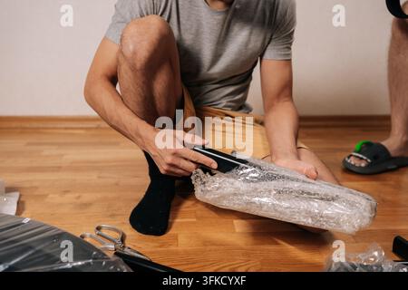 Man unpackaging new furniture components from box wrapped in bubble wrap, preparing to assemble office chair for his personal workspace or home office Stock Photo