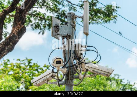 A cluster of outdoor surveillance cameras and wireless equipment is mounted on a metal pole against trees and blue sky. Stock Photo