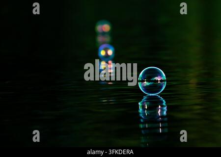 A single luminous bubble balances on dark green water with a glowing reflection. Minimal surroundings and deep tones emphasize its fragile form and qu Stock Photo