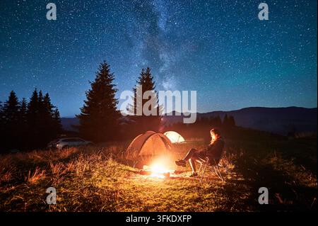 Hiker woman relaxes by campfire, illuminated by its warm glow, with tent nearby under stunning, star-filled sky. Milky Way stretches overhead, adding magical touch to serene mountain setting. Stock Photo