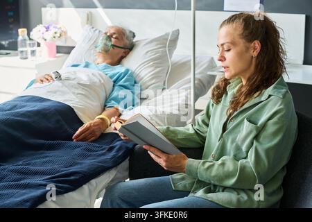 Woman Reading Book to Senior Hospital Patient Wearing Oxygen Mask Stock Photo