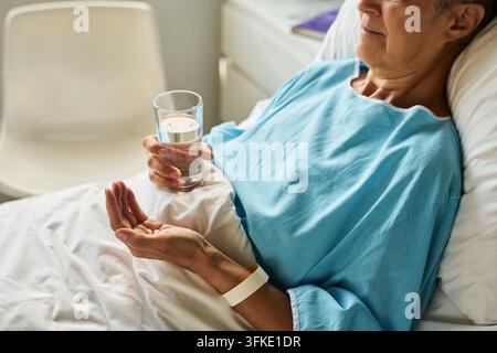 Senior Woman Taking Medication with Glass of Water in Hospital Bed Stock Photo