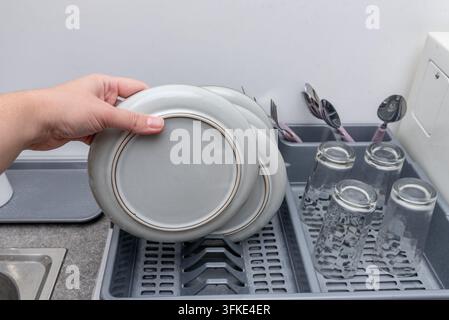 Person places dinner plates in dishwasher rack, completing household chore. Hand steadies each ceramic dish beside drinking glasses and silverware, pr Stock Photo