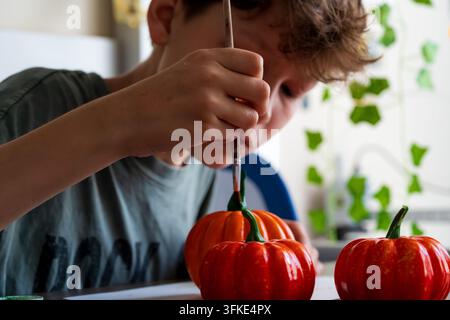 Boy painting a 3D printed pumpkin with a brush. Kid decorating Halloween craft with green stem. Autumn DIY activity and creative hobby. Stock Photo