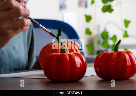 Boy painting a 3D printed pumpkin with a brush. Kid decorating Halloween craft with green stem. Autumn DIY activity and creative hobby. Stock Photo