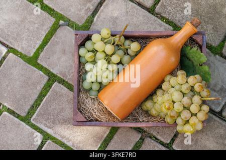 Overhead view of a corked ceramic bottle, green grapes and vine leaves in a wooden crate on paving stones, with copy space on the left. Stock Photo