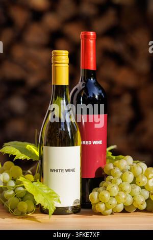 White and red wine bottles arranged with green grape bunches and vine leaves on a wooden table, against a dark, softly blurred firewood background. Stock Photo