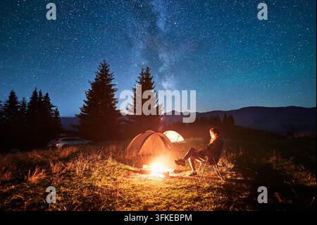 Hiker woman relaxes by campfire, illuminated by its warm glow, with tent nearby under stunning, star-filled sky. Milky Way stretches overhead, adding Stock Photo