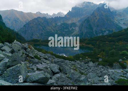 A tranquil alpine lake reflects towering jagged mountain peaks under a cloudy sky, surrounded by rocky terrain and green pine trees. Stock Photo