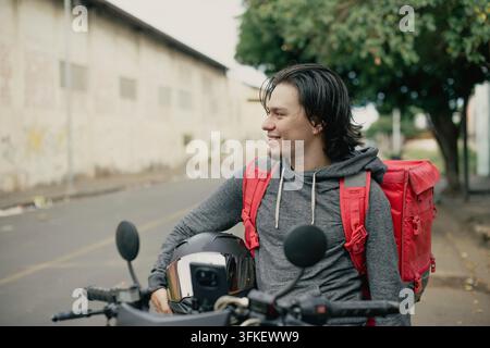 Young delivery courier with red insulated backpack and helmet by motorcycle. Smiling delivery man holding motorcycle helmet with red thermal backpack Stock Photo