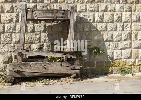 A weathered wooden structure stands against a textured stone wall beside a paved walkway. Strong sunlight creates defined shadows across the rough woo Stock Photo