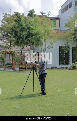An Asian photographer operates a professional mirrorless camera with a telephoto lens on a grassy lawn at the outdoor noon time. Stock Photo