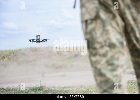 FPV quadcopter drone flies low above a dusty training field after takeoff while a person in camouflage clothing stands in the foreground. Dust rises f Stock Photo