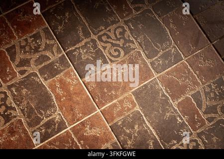 Close-up of rustic brown ceramic floor tiles featuring decorative patterns, visible grout lines and worn surface texture. Ideal for interior design, f Stock Photo