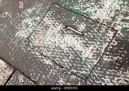 Close-up of a weathered metal manhole cover showing diamond plate pattern, chipped white and green paint, rusted handle and gritty urban texture, suit Stock Photo
