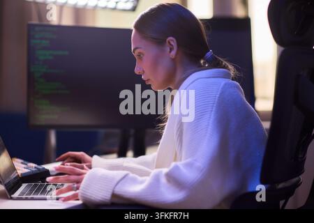 Female coder focused. Young professional working late at computer under soft lighting. Dedicated woman coding intensely at desk with monitor showing Stock Photo