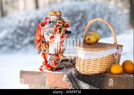 Winter still life featuring a traditional metal samovar with bagels, fresh apples, oranges and a wicker basket outdoors. Cozy rustic composition symbo Stock Photo