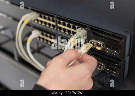 A male IT technician in his 30s works in a data center, connecting Ethernet cables to server hardware. The scene highlights hardware maintenance, netw Stock Photo