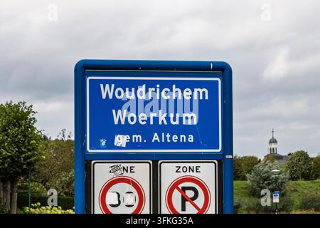 Woudrichem, Netherlands: Official blue town entry sign reading Woudrichem and local name Woerkum, with traffic signs and a blurred church tower in the Stock Photo