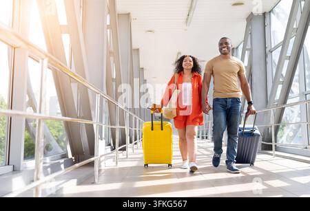 Happy young Black couple walking hand in hand through sunlit airport corridor Stock Photo