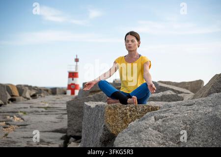 A middle-aged woman sits in a meditative lotus pose on large grey boulders by the sea during the late afternoon. The scene features a red and white Stock Photo