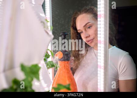 Woman refreshing mirror with spray. Casual woman cleaning mirror with spray in bright room. Relaxed woman using spray to clean mirror on vanity during Stock Photo