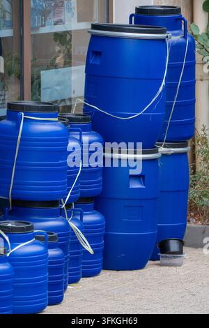 A tall stack of large blue plastic barrels and chemical storage drums tied together with white string stands outdoors on a paved surface outside a bui Stock Photo