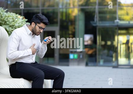 Young man experiencing a sudden asthma attack while sitting on a public bench, holding his chest and using a blue portable inhaler for respiratory relief and managing his breathlessness outdoors Stock Photo